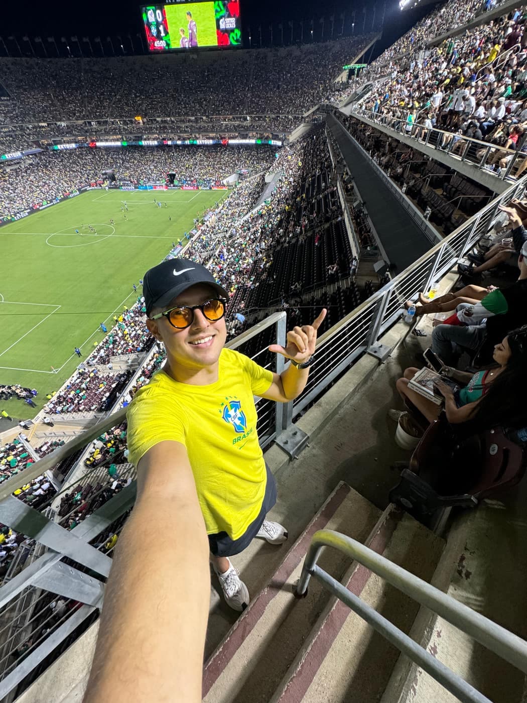 Selfie do Thiago à noite em um estádio de futebol lotado, sorrindo, de óculos de lente âmbar